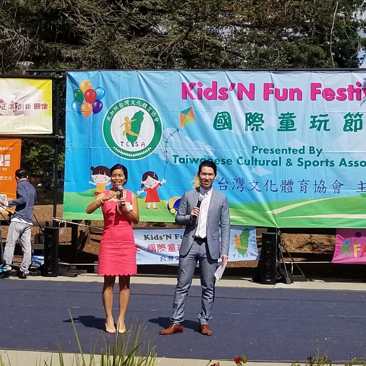 Two people with microphones on an outdoor stage in front of a Kids'N Fun Festival banner.