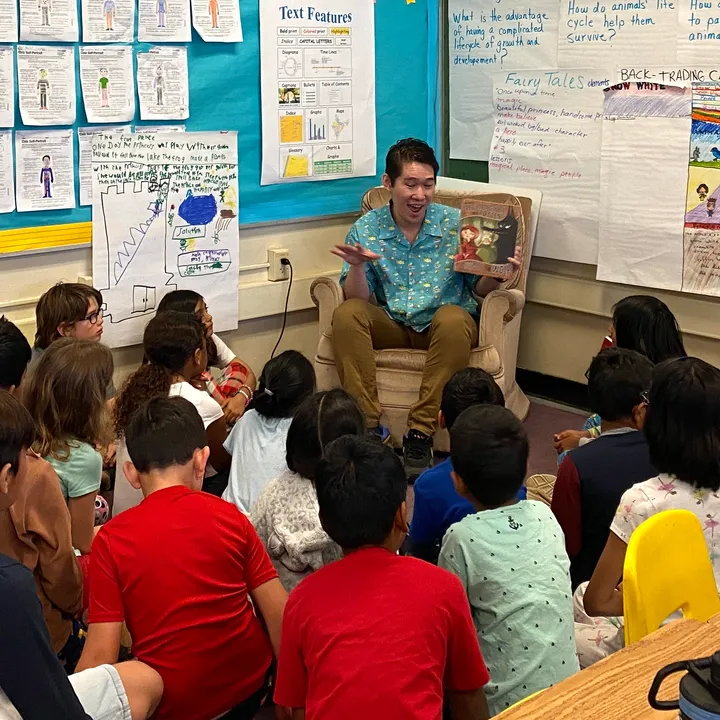 Reading a picture book aloud from an armchair to a class of children sitting on a classroom floor.