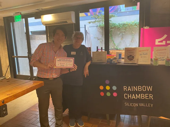 Two people at a check-in table beside a Rainbow Chamber Silicon Valley banner, one holding a card for the Silicon Valley Pride kickoff mixer.