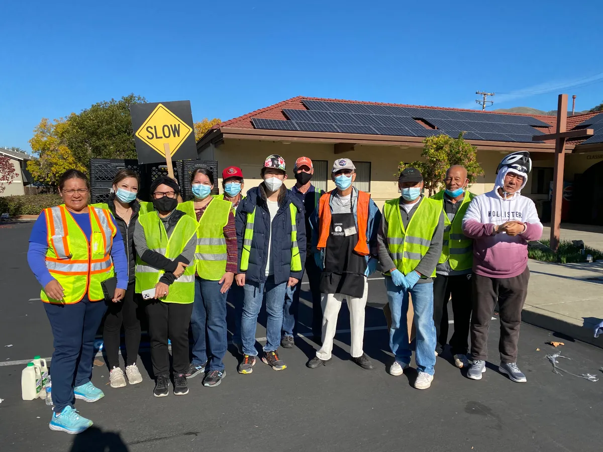 A line of masked volunteers in high-visibility vests in a car park, one wearing a Second Harvest of Silicon Valley apron.