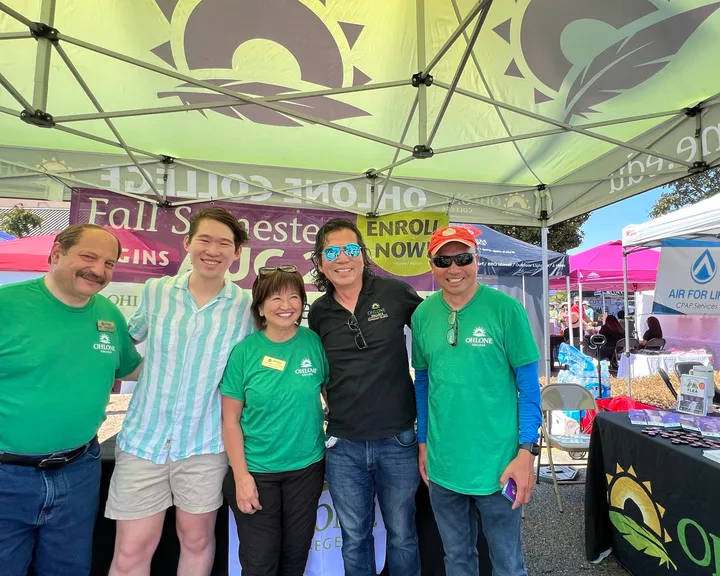 Five people in Ohlone College T-shirts under the college's canopy at an outdoor festival, in front of a banner reading Fall Semester Begins.